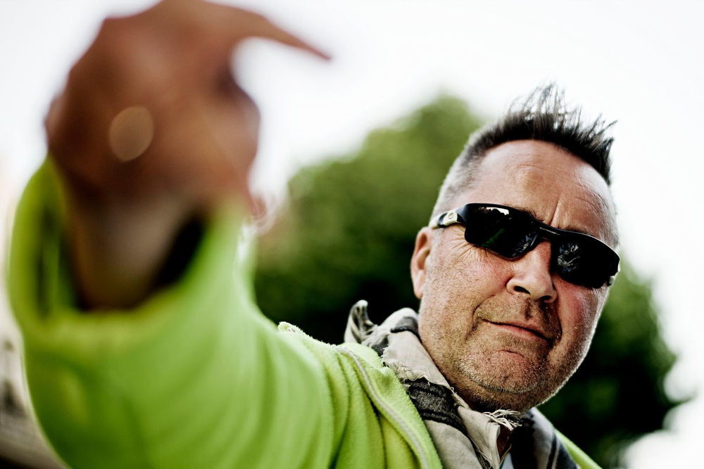 Nigel Kennedy – zdjęcie mężczyzny w średnim wieku w okularach przeciwsłonecznych, zielonej bluzie ,na twarzy lekki zarost, wokół szyi owinięty lekki szal lub apaszka. Artysta wyciąga w kierunku obiektywu prawą rękę. Zdjęcie w plenerze – w tle widać nieostre korony drzew.