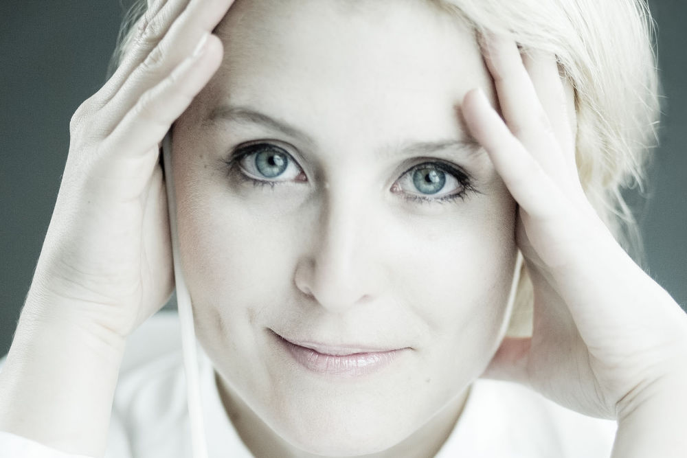 Portret artystki – Patrycji Pieczary. Ciasny kadr przedstawiający lekko uśmiechniętą twarz kobiety podpartą w okolicach skroni dłońmi. Niebieskie oczy i blond włosy.