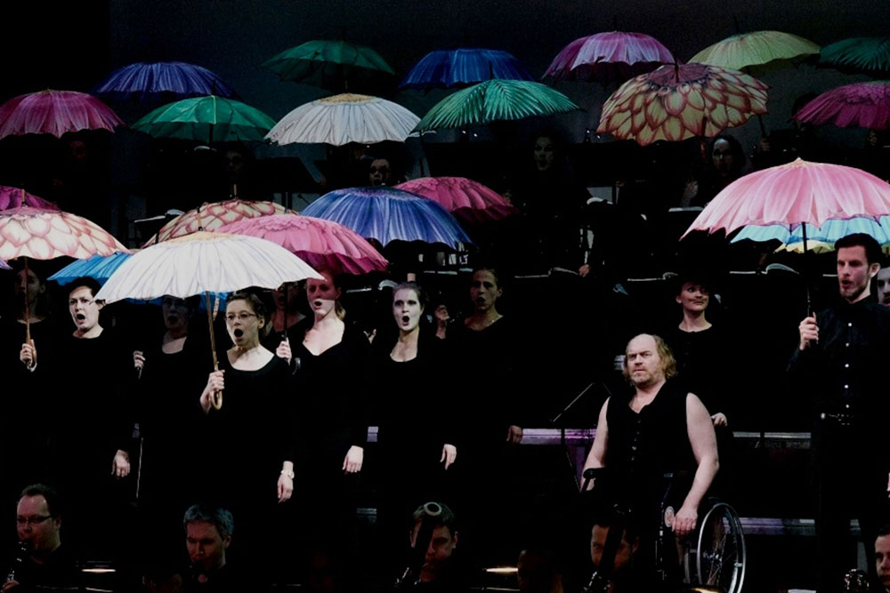 Na zdjęciu artyści chóru w czarnych strojach. Śpiewacy trzymają nad głowami rozłożone kolorowe parasole. Jeden z artystów na wózku inwalidzkim.