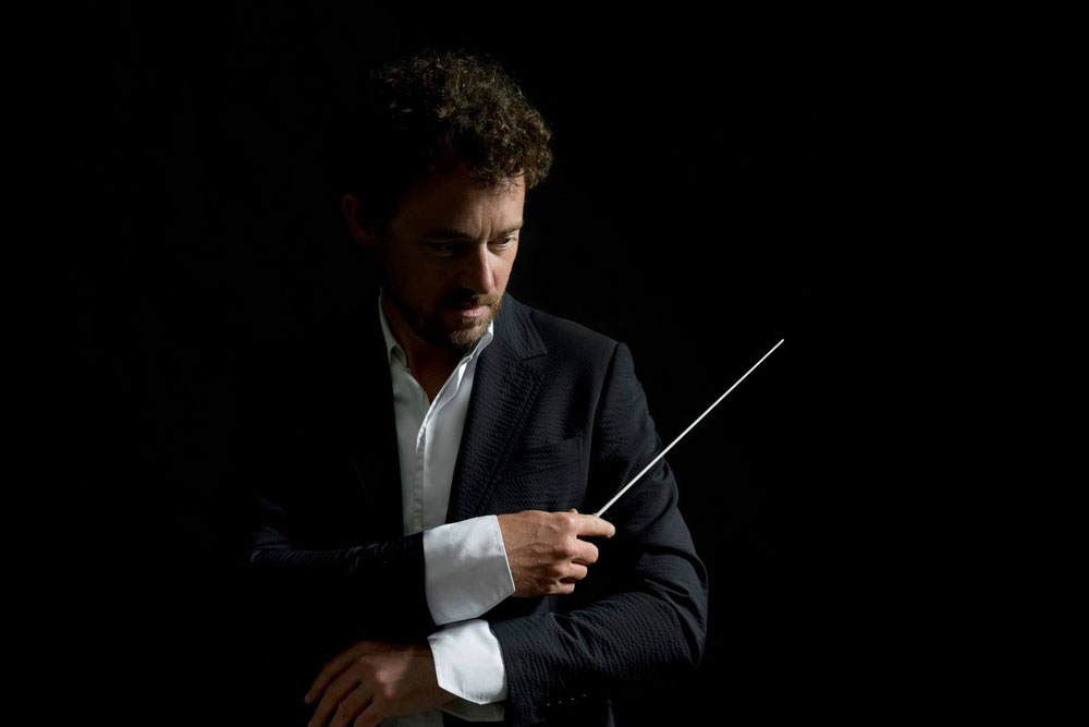 Na zdjęciu Jean-Luc Tingaud. Portret dyrygenta wyłaniającego się z półmroku. Światło pada na lewy profil twarzy. Mężczyzna w czarnym smokingu i białej koszuli z szerokimi mankietami. Skrzyżowane ręce trzymane blisko tułowia. W prawej dłoni batuta.