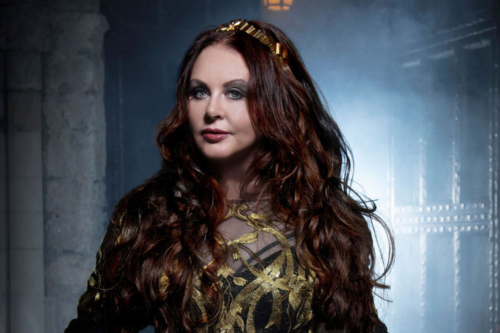 Na zdjęciu Sara Bbrightman. Na pierwszym planie portret artystki patrzącej na odbiorcę. Rozpuszczone, długie, brązowe włosy przewiązane złotą wstążką odsłaniającą się w górnej części głowy. Widoczna górna część garderoby – najpewniej czarnej sukni z przeźroczystym dekoltem ozdobionej ażurowymi roślinnymi elementami w kolorze złotym. W tle przebija się rustykalna architektura w niebieskich odcieniach