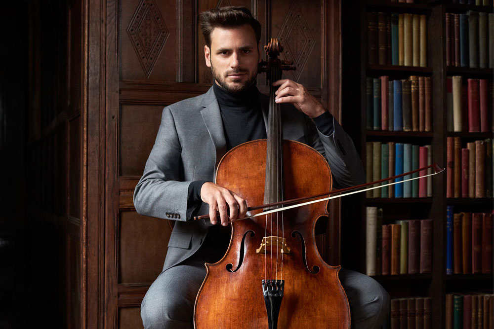 Na zdjęciu Stjepan Hauser. Mężczyzna w pozycji siedzącej z wiolonczelą. W prawej ręce smyczek skrzyżowany z instrumentem. Prawa ręka na gryfie wiolonczeli. Muzyk w stalowym garniturze i czarnym golfie. W tle drewniana biblioteka.