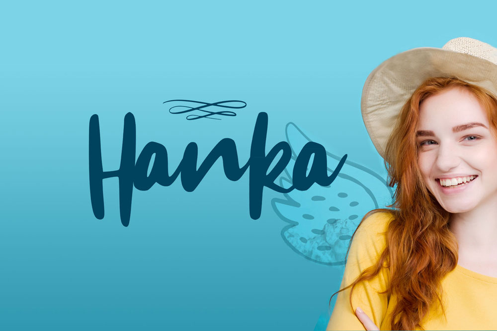 Na zdjęciu uśmiechnięta, rudowłosa, młoda dziewczyna w słomkowym kapeluszu i żółtej bluzce. Dziewczyna ma dorysowane kreską skrzydło, jakby anielskieObok Stylizowany na odręczny napis "Hanka". Całość na błękitnym tle. Obraz emanuje radością i optymizmem
