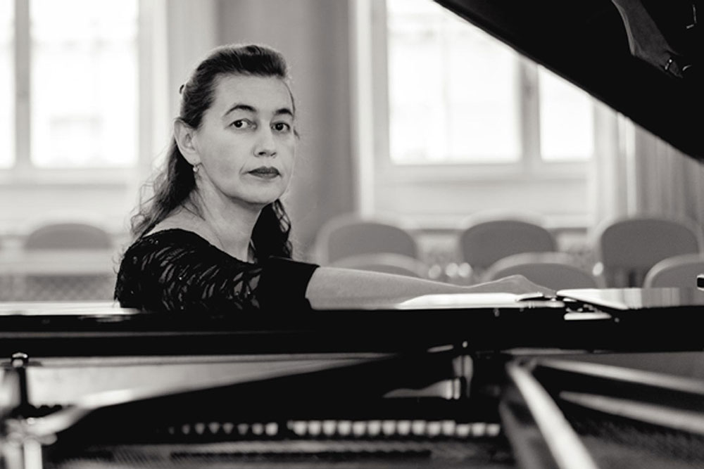 Czarno-białe pozowane zdjęcie przedstawia pianistkę Lilyę Zilberstein siedzącą przy fortepianie. Prawa ręka artystki spoczywa na złożonym pulpicie instrumentu.