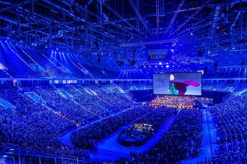 Na zdjęciu wnętrze Tauron Areny wypełnionej widzami. W oddali Ekran kinowy , ze sceną z filmu Kraina Lodu. Zdjęcie w odcieniu atramentowego światła.