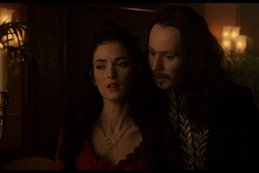 Na zdjęciu kadr z filmu "Drakula" z tysiąc dziewięćset dziewięćdziesiątego drugiego roku. Na pierwszym planie kobieta i stojący za nią mężczyzna szepczący jej do ucha. Scena w półmroku. Postaci w strojach z epoki.