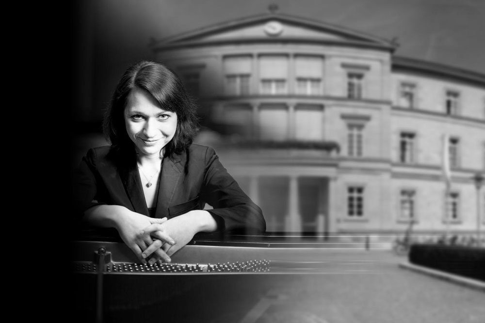Czarno-biały kolarz. Po lewej uśmiechnięta pianistka Anastasia Voltchok siedzącą przy fortepianie. Patrząca w obiektyw artystka trzyma dłonie ze skrzyżowanymi palcami na kluczach strun fortepianu. Portret z prawej strony przechodzący w zdjęcie budynku uniwersytetu w Tübingen