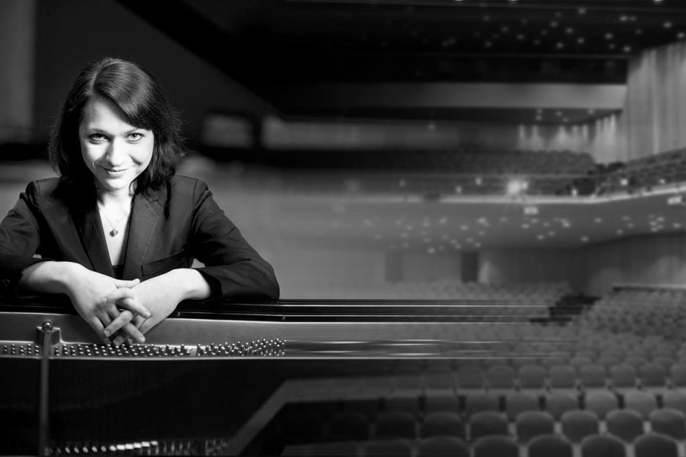 Czarno-biały kolarz. Po lewej uśmiechnięta pianistka Anastasia Voltchok siedzącą przy fortepianie. Patrząca w obiektyw artystka trzyma dłonie ze skrzyżowanymi palcami na kluczach strun fortepianu. Portret z prawej strony przechodzący w zdjęcie sali Congress Center.